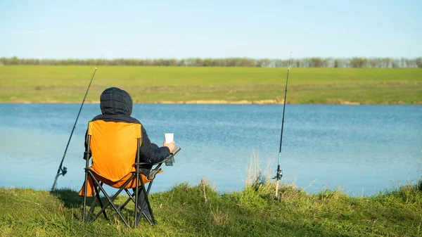 A man on fishing sitting in a chair and holding a cup, two fishing rods on stands, water on the background, good weather