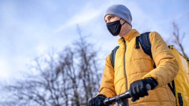 Scooterlı yemek teslimatçısı. Siyah tıbbi maske, sarı sırt çantası ve ceket. Kış, arka planda ağaçlar