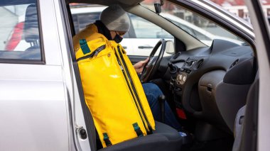 Arabadaki siyah tıbbi maskeli kurye telefonunda, teslimat çantası da koltukta. Yemek dağıtım servisi.