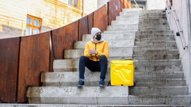 Kışın teslimatçı merdivenlerde oturan sarı sırt çantalı adam.