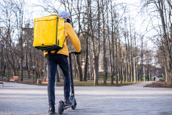 Food delivery man on a scooter in a park. Black medical mask, yellow backpack and jacket. Winter
