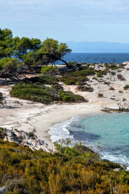 Çevresinde yeşillikler, kayalar, çalılar ve ağaçlar, dalgalı mavi sular, Yunanistan