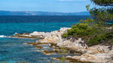 Çevresinde yeşillikler, kayalar, çalılar ve ağaçlar, dalgalı mavi sular, Yunanistan