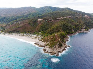 Sahile yakın kamp yapan Ege kıyıları, etrafındaki yeşillikler, kayalar, çalılar ve ağaçlar, arka planda tepeler, dalgalı mavi sular, insansız hava aracı Yunanistan 'ın manzarası