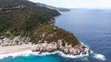 Sahile yakın kamp yapan Ege kıyıları, etrafındaki yeşillikler, kayalar, çalılar ve ağaçlar, arka planda tepeler, dalgalı mavi sular, insansız hava aracı Yunanistan 'ın manzarası