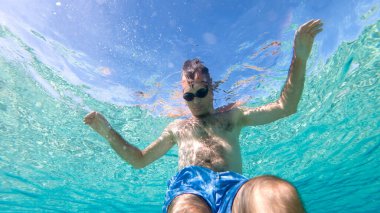 Yüzme gözlükleriyle suyun altında yüzen beyaz bir adam. Altında kamera var. Mavi şeffaf su.