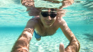 Yüzme gözlükleriyle suyun altında yüzen beyaz bir adam elinde kamera, mavi şeffaf su.