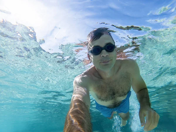 Caucasian man swimming under the water in swimming goggles holding a camera, blue transparent water