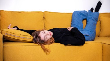 Siyah kazaklı ve kot pantolonlu genç bir kızıl evde sarı bir kanepede uyuyor.