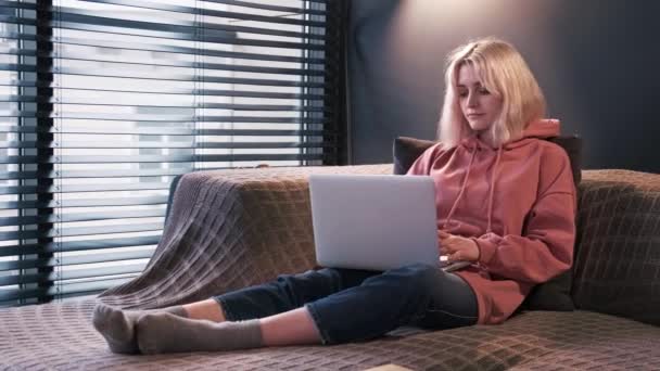 若いコンテンツ作成者のブロンドの女の子は 窓の近くのソファに座って彼女のラップトップにあります 家で働くこと — ストック動画