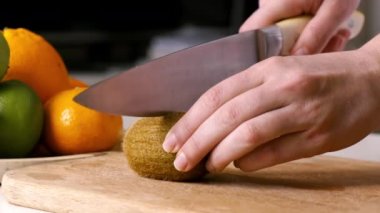 Tahta bir tahtanın üzerinde kivi dilimleyen bir kadın. Arka planda meyveler var. Yavaş çekim