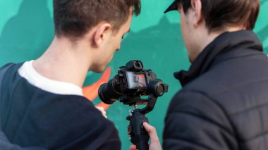 İki beyaz adam kamera ekranına bakıyor. Sabit, renkli bir arka plan.