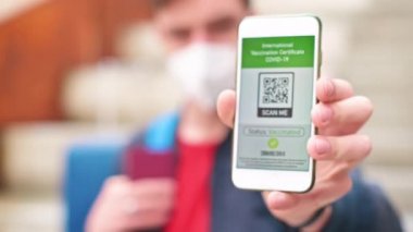 Tıbbi maskeli bir adam akıllı telefondaki Uluslararası Aşı Sertifikası COVID-19 QR kodunu gösteriyor