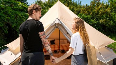 Glamping 'de çadırın yanında el ele tutuşan genç bir çift. Çevrede yeşillik