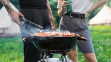 Genç bir çift ızgarada et pişiriyor. Çevrede yeşillik. Glamping