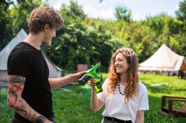 Bira içen, gülümseyen, şişe tokuşturan genç bir çift. Çevrede yeşillik. Glamping