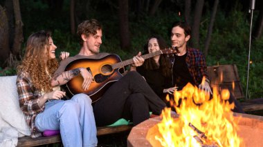 Glamping 'deki kamp ateşinin yanında bir grup mutlu genç arkadaş. İki kadın ve erkek. Gitar çalmak