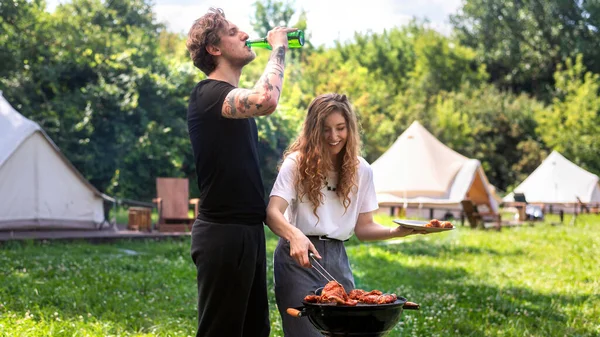 Young couple frying meat on the grill and drinking beer. Greenery around. Glamping