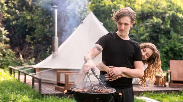 Young hugging couple frying meat on the grill. Greenery around. Glamping