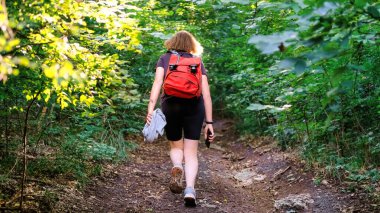 Turuncu sırt çantalı kızıl saçlı kadın ormanda bir patikada yürüyor, yeşillik içinde.