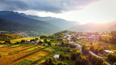 Gün batımında Gürcistan 'da bir köyün insansız hava aracı görüntüsü. Vadi, tarlalar ve binalar, dağlar ve tepeler yeşillikle kaplı, bulutlu gökyüzü