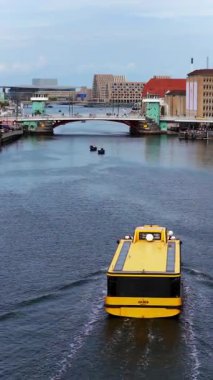 Kopenhag, Danimarka - 4 Ağustos 2025: Arkasında köprüler ve şehir mimarisi bulunan Kopenhag limanı boyunca giden sarı bir liman otobüsü feribotunun insansız hava aracı görüntüsü. Dikey