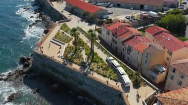 Antibes Eski Şehir surları boyunca yürüyen insanların hava aracı görüntüsü, palmiye ağaçları, peyzaj bahçeleri ve kayalık kıyılara çarpan dalgalar.