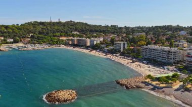 Antibes kumlu plajının hava aracı görüntüsü güneşlenenlerle ve yüzücülerle dolu, deniz manzaralı apartmanlar ve arka planda yeşil tepeler var.
