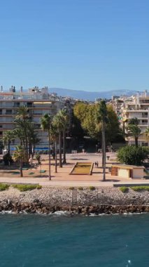 Antibes sahil şeridinin hava aracı görüntüsü palmiye ağaçları ve modern dairelerle çevrili, Akdeniz ve arka planda uzak dağlar. Dikey