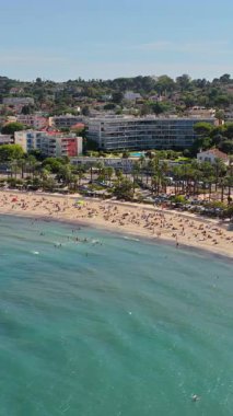 Antibes plajı ve gezinti güvertesi, palmiye ağaçları ve dairelerle kaplı, turkuaz Akdeniz suları ve körfezdeki yüzücülerle dolu. Dikey