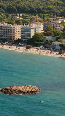 Antibes kumlu plajının deniz drone görüntüsü güneşlenenlerle ve yüzücülerle, deniz kenarındaki apartmanlarla ve arka planda yeşil tepelerle dolu. Dikey