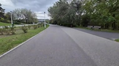 Boş bir park yolunun geniş görünümü bulutlu bir sonbahar gününde sıralanmış banklar ve yeşil ağaçlarla dolu.