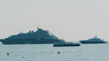 Büyük yolcu gemisi ve bulutlu gökyüzünün altında durgun mavi denize demir atmış lüks yatlar.