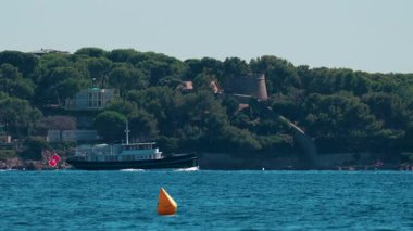 Fransız Rivierası 'ndaki açık mavi gökyüzünün altında, çam ağaçları ve villalarla çevrili Cannes kıyısı boyunca zarif siyah bir yat yelken açıyor.