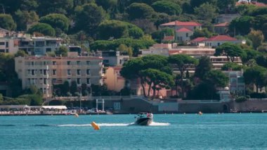 Zarif bir motorlu tekne, arka planda bereketli ağaçlar ve kıyı binaları olan berrak mavi sularda hızla ilerliyor.