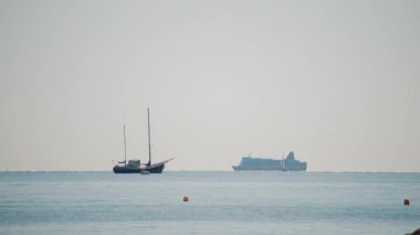 Sakin bir denizin geniş bir görüntüsü, yelkenli ve açık mavi gökyüzünün altında yelken açan büyük bir yolcu gemisi.