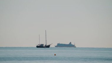 Sakin bir denizin geniş bir görüntüsü, yelkenli ve açık mavi gökyüzünün altında yelken açan büyük bir yolcu gemisi.