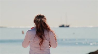 Güneş gözlüğü takan, gülümseyen bir kadın ve beyaz bir gömlek güneşin altında, arkasında deniz parıldıyor.