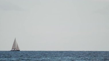 Beyaz bir yelkenli, açık güneşli bir günde mavi dalgalarda yol alır. Ufuk arkalarda uzanıp durur.