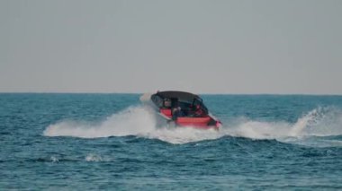 Siyah bir gölgeliği ve kırmızı iç yüzeyi olan lüks bir tekne açık denizde hızla ilerlerken arkasında beyaz dalgalardan oluşan bir iz bırakıyor.