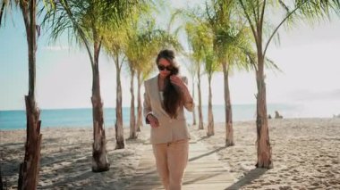 Bej takım elbiseli şık bir kadın ve güneş gözlüğü sakince yürüyor palmiye ağacından bir patika boyunca denize doğru