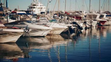 Açık mavi gökyüzünün altında güneşli bir marinada demirlemiş yat ve tekneleri görüyorum.