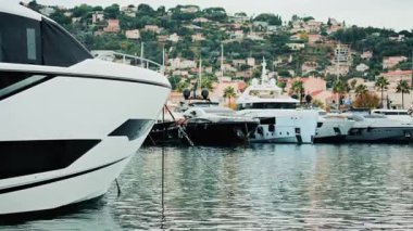 Lüks yatlar sakin bir marinaya demirlemiş ve arka planda manzaralı bir yamaç kasabası var.