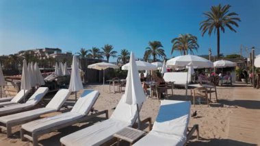 Cannes, Fransa - 3 Ekim 2025: Turistler güneşleniyor ve kumlu bir sahilde, palmiye ağaçları ve mavi gökyüzü olan beyaz şemsiyeler altında kitap okuyorlar