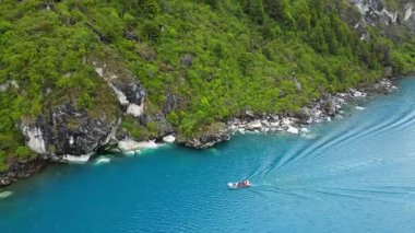 Patagonya 'daki orman kayalıklarıyla çevrili turkuaz bir gölde süzülen küçük bir teknenin hava aracı görüntüsü.