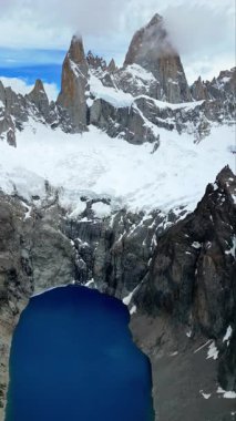 Fitz Roy Dağı 'nın görkemli zirvelerinin hava aracı görüntüsü kayalık kayalıklarla çevrili derin mavi bir buzul gölü üzerinde yükseliyor. Dikey