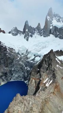 Fitz Roy 'un, kayalık kayalıklar ve kar yamaçları tarafından çerçevelenmiş koyu mavi bir buzul gölü üzerindeki gökyüzü drone görüntüsü. Dikey
