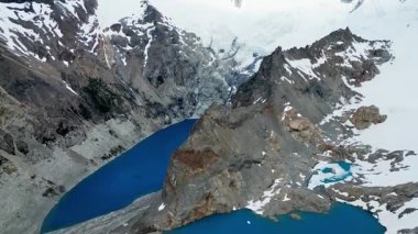 İki derin mavi buzul gölünün hava aracı görüntüsü kayalık tepelerle ayrılmış ve karla kaplı dağlarla ağır çekimde çevrili.