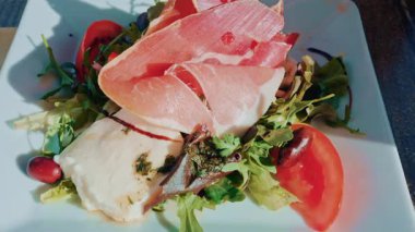 Taze mozarella, taze et dilimleri, domatesler, zeytinler ve karışık yeşillikleri beyaz bir tabakta güpegündüz servis et.