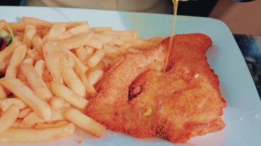 Mavi bir tabakta altın patates kızartmasıyla servis edilen kızarmış fileto, üzerine zeytinyağı dökülen bir el gibi.